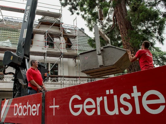 Arbeiter montieren ein Baugerüst an einem Hausdach mit Kran und Gerüstbau-Fahrzeug.