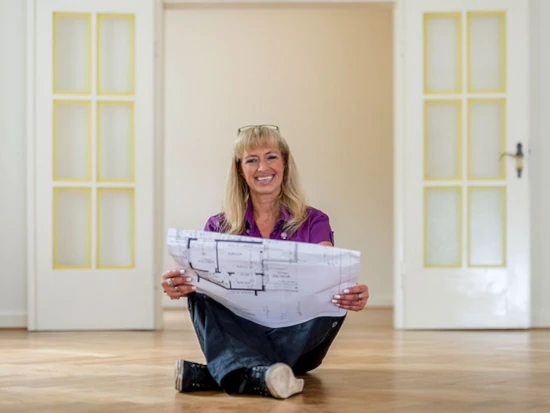 Frau sitzt auf Parkettboden und hält Bauplan in heller, renovierter Wohnung in den Händen.