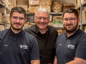 Drei Männer stehen lächelnd in einem Lager zwischen Regalen mit Verpackungsmaterial.