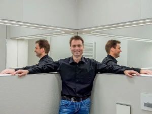 Mann steht in modernem Badezimmer mit Spiegelwand, trägt schwarze Bluse und blaue Jeans.
