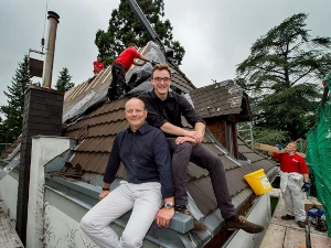 Zwei Männer sitzen auf einem Hausdach, im Hintergrund arbeiten Handwerker an der Dacheindeckung.