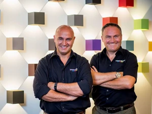 Zwei lächelnde Männer stehen vor einer farbenfrohen Wand mit modernen Lampen-Designs.