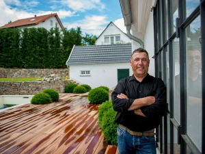Mann steht lächelnd auf moderner Holzterrasse vor Haus mit Garten und Glasfassade.