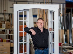 Ein Schreiner steht in einer Werkstatt hinter einem weißen Holzfenster mit Sprossen.