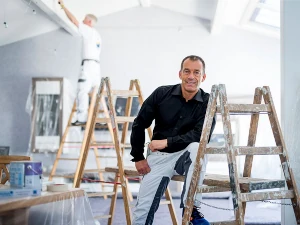 Zwei Maler renovieren einen hellen Innenraum mit Leitern und Malerzubehör.