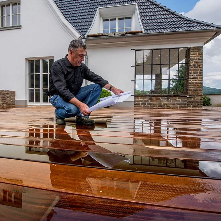 Ein Mann prüft Baupläne auf einer nassen Holzterrasse vor einem modernen Haus.