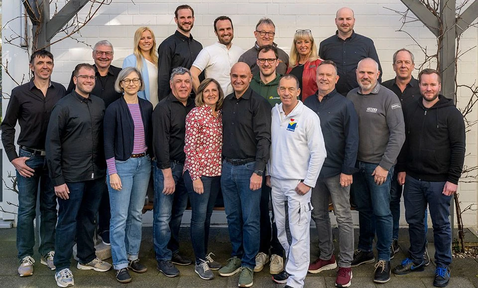 Gruppenfoto von lächelnden Mitarbeiterinnen und Mitarbeitern vor einer weißen Wand draußen.